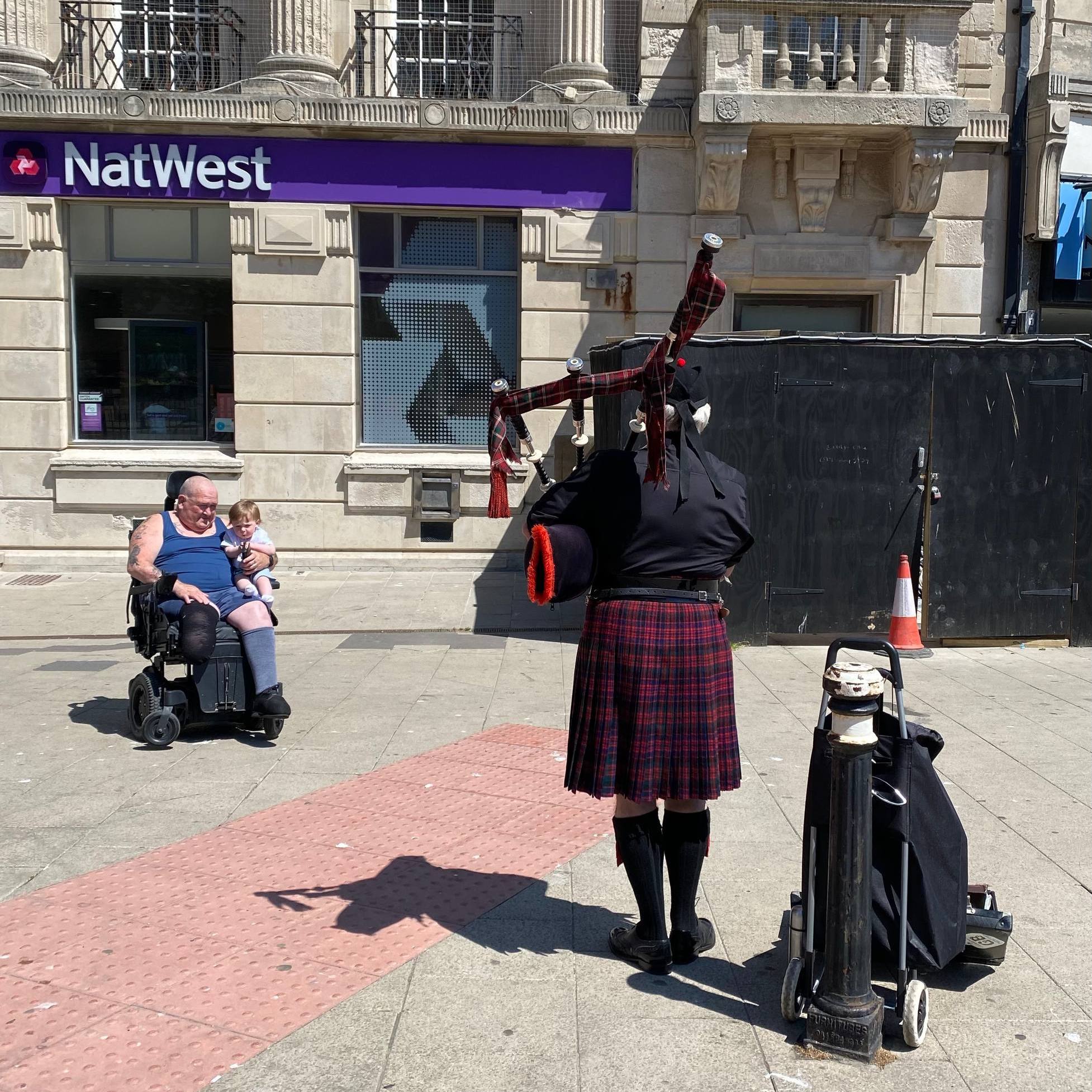Man in wheelchair with one leg, holding an small baby in his lap while listening to a bagpipe player with his back turned to camera. 